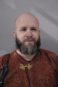 Close-up of Society Chatelaine, Andrew Harness. He is bald, with a beard and mustache, and wearing a maroon Persian style tunic with a gold closure.